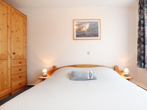 Ein Schlafzimmer mit einem Holzschrank, einem Bett mit weißer Matratze und blauem Kissen, zwei Nachttischlampen und einem gerahmten Bild von einem Vogel an der Wand.