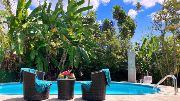 Area piscina con sedie in vimini e un tavolo, immersa in rigogliosa vegetazione tropicale.