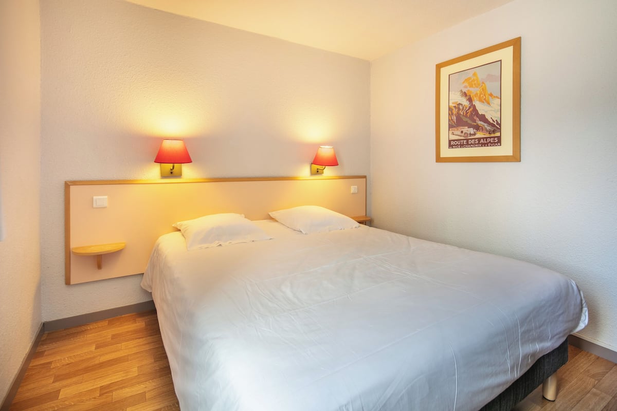 Ein ordentlich bezogenes Bett mit weißen Leinen, zwei Nachttische mit Lampen, ein kleines Regal und ein gerahmtes Bild an der Wand.