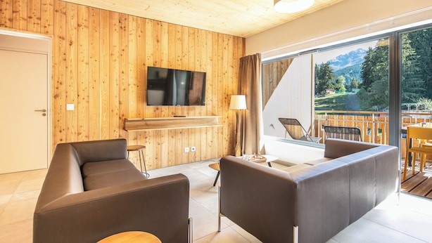 A modern living room with wooden panel walls, a flat-screen TV, a sofa, a coffee table, and a balcony with a view of mountains. - VVF Les 7 Laux Massif de Belledonne, Prapoutel (Les Adrets)