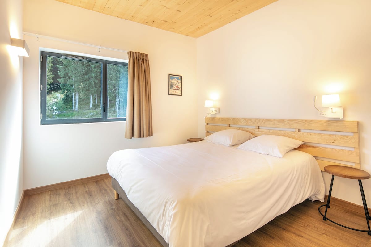 Ein Schlafzimmer mit einem hölzernen Kopfende, einem großen Bett, einem Nachttisch, Vorhängen an einem Fenster und einem gerahmten Bild an der Wand.