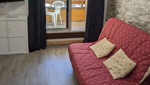 A room with a red sofa, two heart-patterned pillows, a TV on the wall, and a view of a balcony with a white chair and a wooden table. - Cozy Studio for 4, Ski-In/Ski-Out, WiFi, Near Shops and Sports Center (Orcières Merlette)