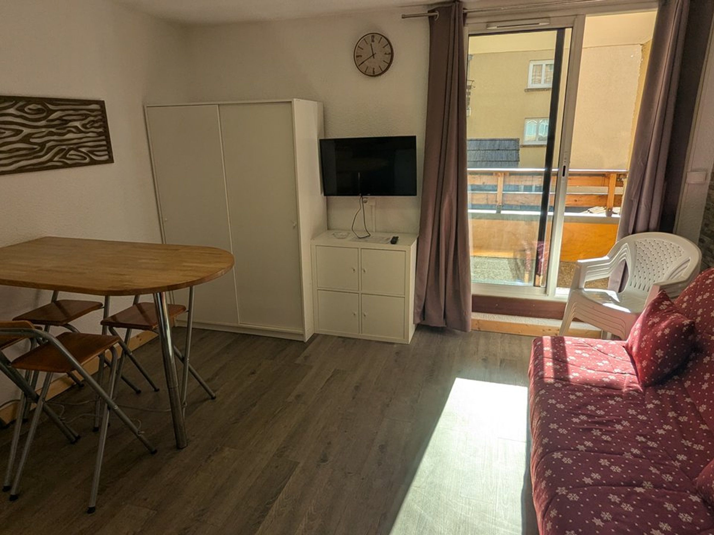 A room with a wooden table, a sofa, a TV on a cabinet, and a sliding door leading to an outdoor area.