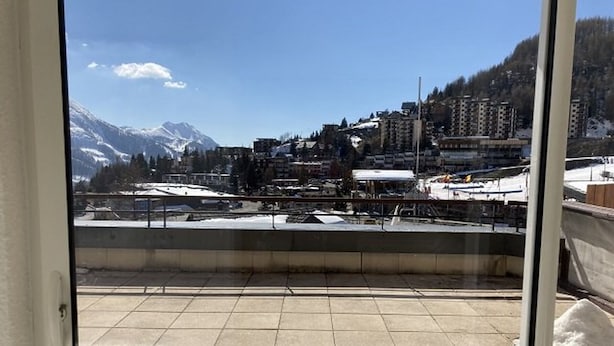 A view from a balcony with tiled flooring, a metal railing, and a snow-covered landscape with buildings and mountains in the background.