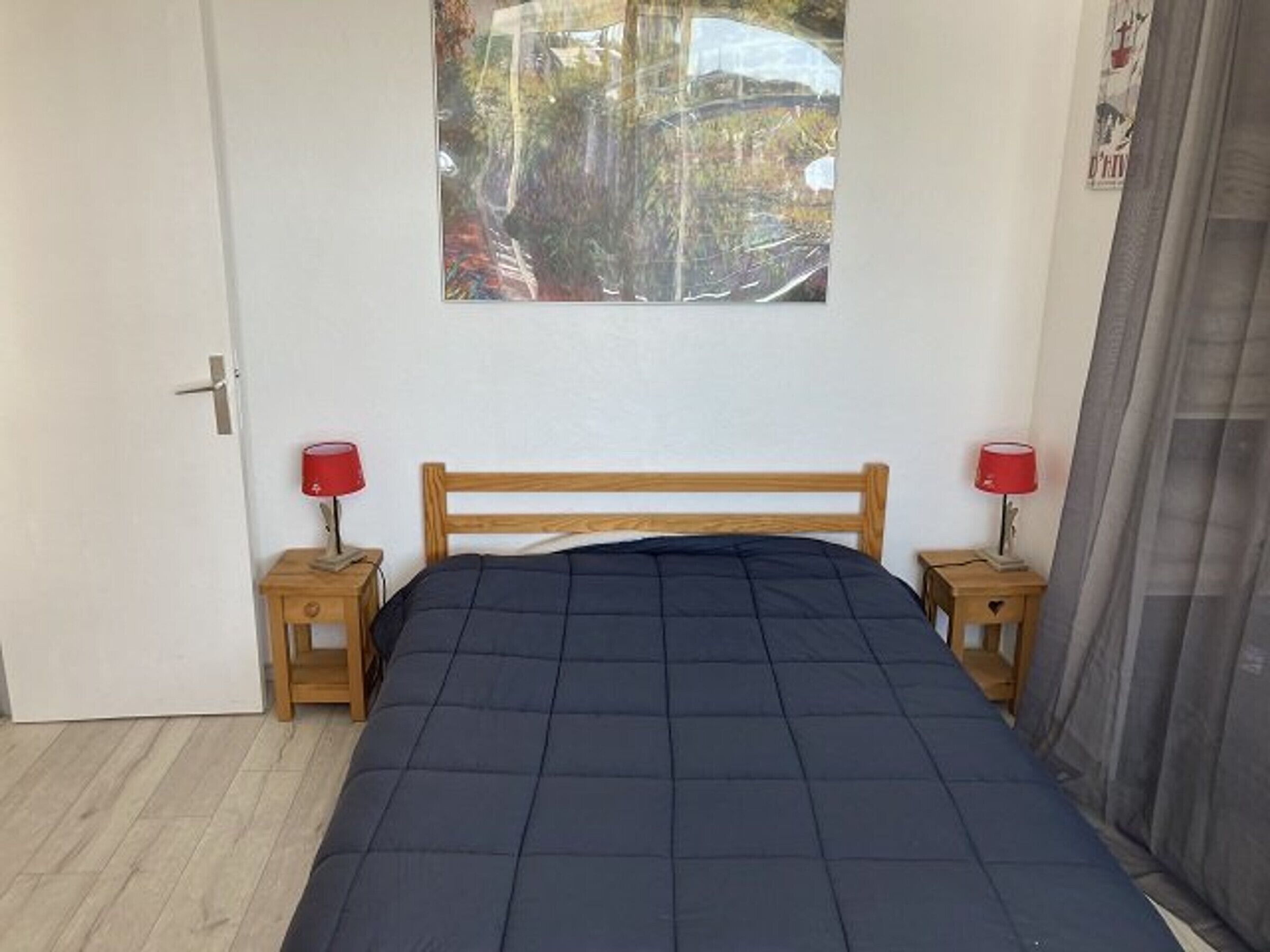 A bedroom with a wooden bed, two bedside tables with red lamps, a door, and a window with a view of trees and a building.