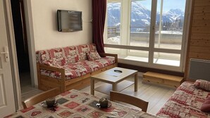 A cozy room with a sofa, a small table, and a bed. There is a TV mounted on the wall and a window with a view of mountains.