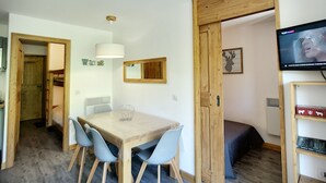 A compact living space with a wooden dining table, two chairs, a TV mounted on the wall, and a bookshelf.