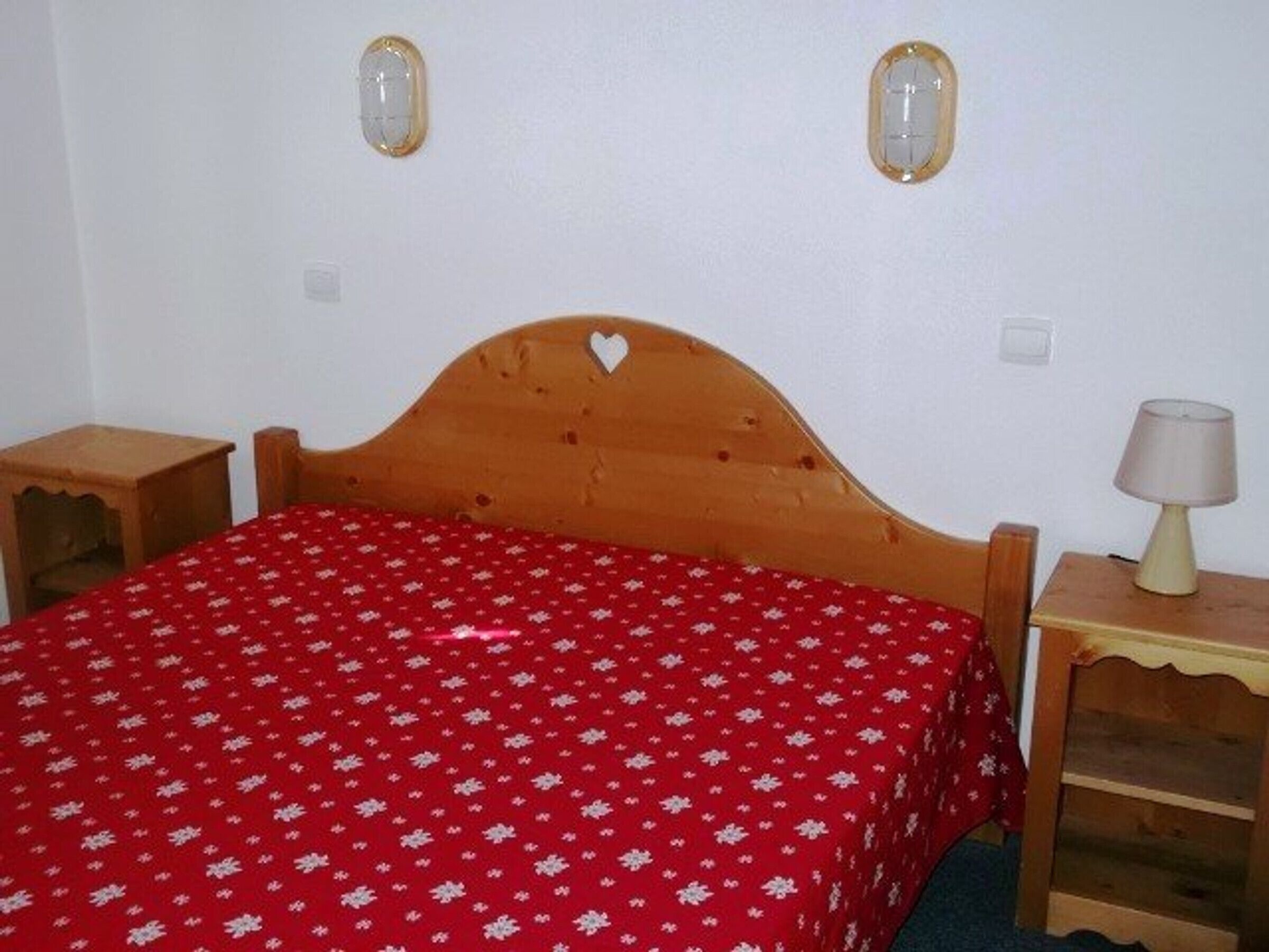 A bedroom with a wooden bed, a red floral bedspread, two bedside tables, and a nightstand with a lamp.
