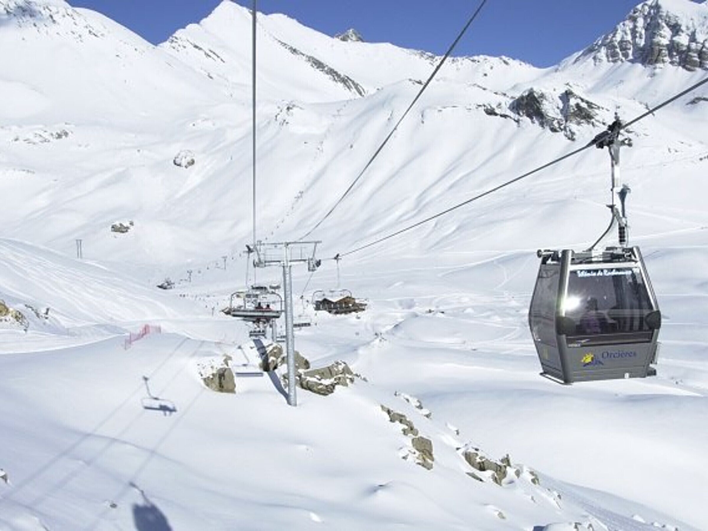 Een stoeltjeslift hangt aan kabels, met een besneeuwd berglandschap op de achtergrond.