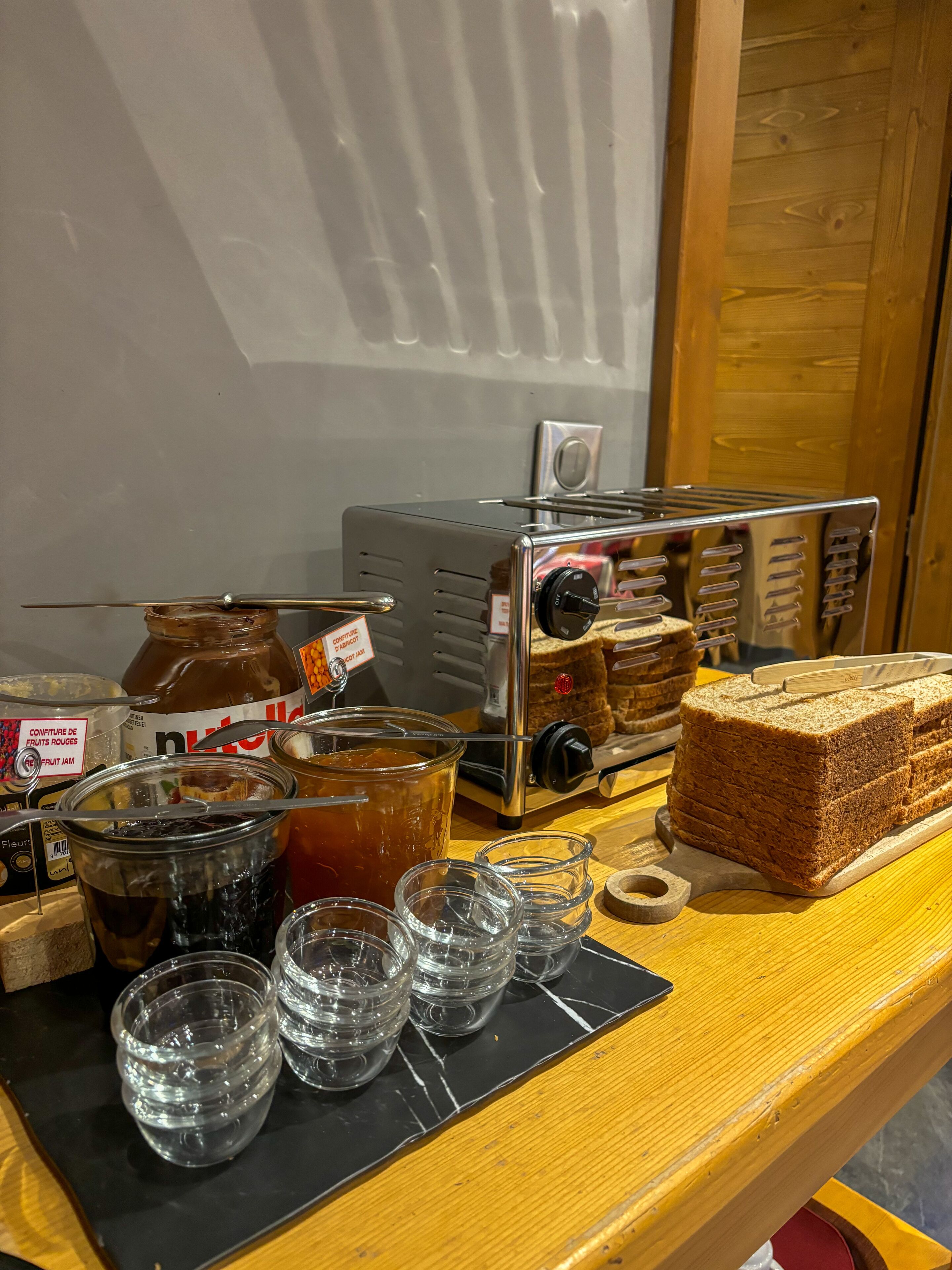 A wooden counter with a toaster, jars of jam, glasses, and a loaf of bread.