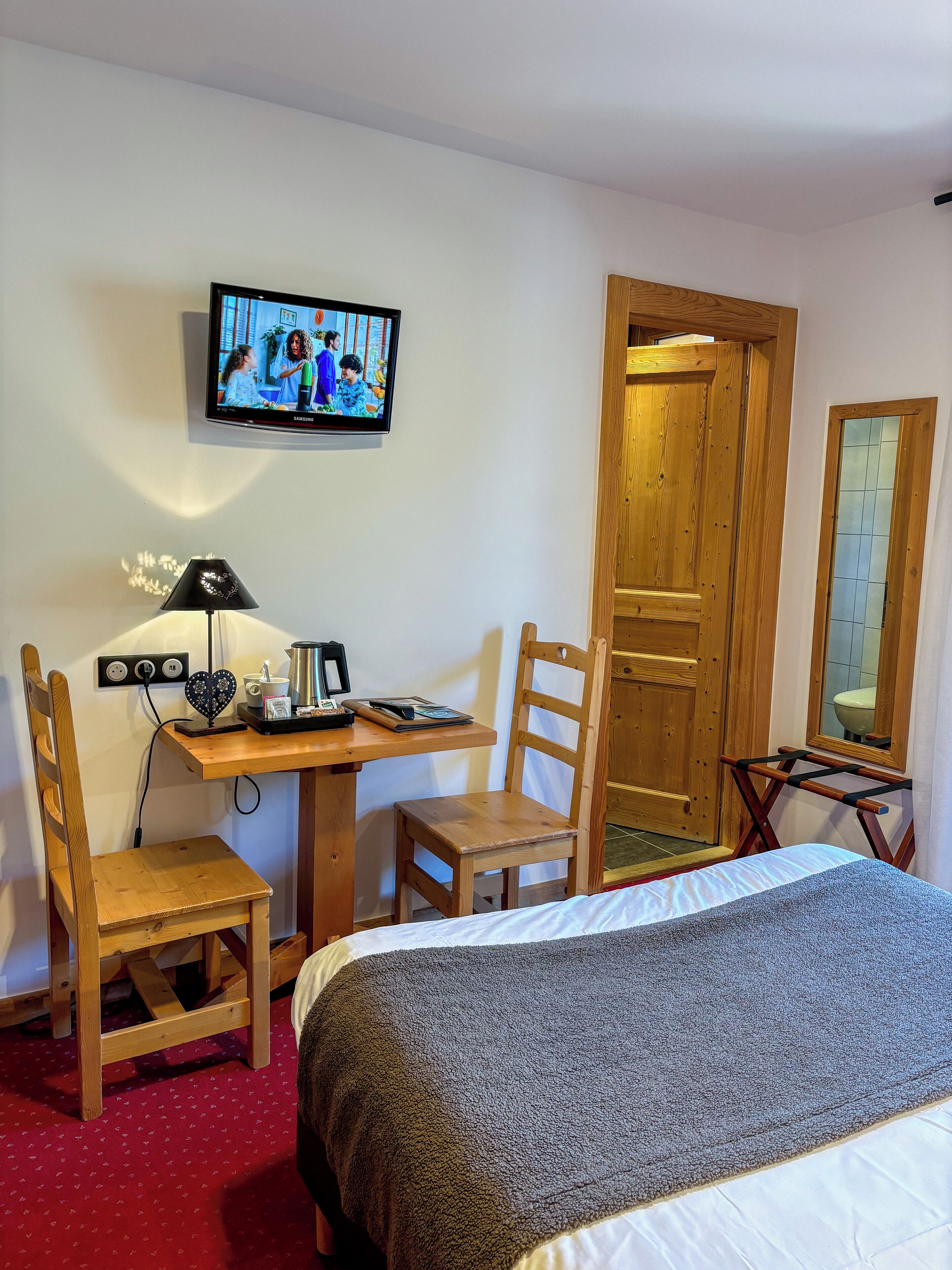 A hotel room with a bed, a desk with a chair, a television, and a wooden door.