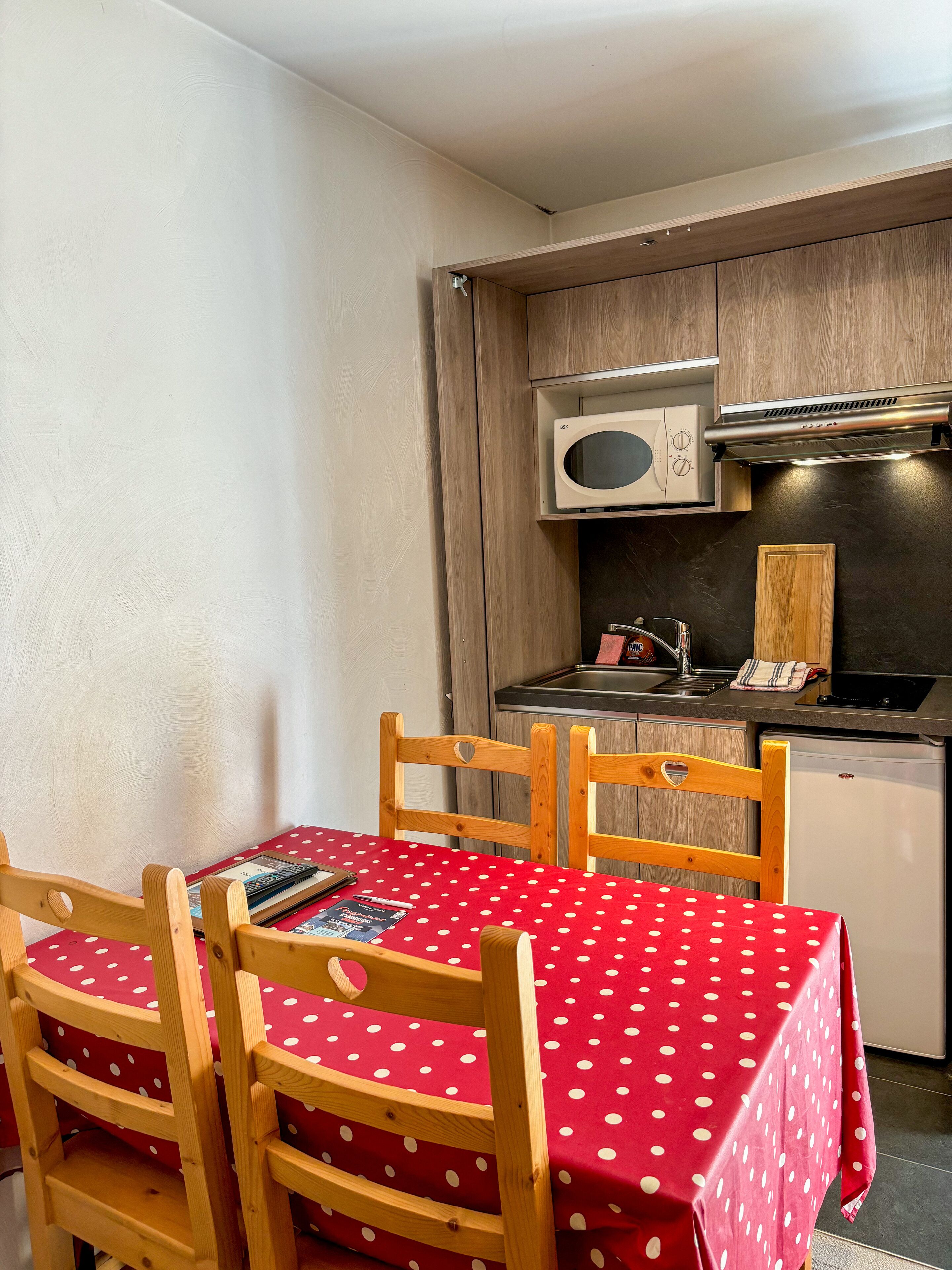 A compact kitchen with a microwave, sink, and a small refrigerator. There is a dining table with a red polka dot tablecloth and wooden chairs.