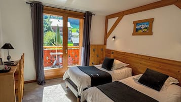 A bedroom with two beds, a desk, and a balcony with a view of a house and greenery.