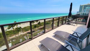 A balcony with a view of the beach and ocean, equipped with lounge chairs and a table.