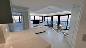 A modern living room with a large window overlooking the sea, a white sofa, and a dining area with chairs and a table.
