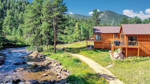 Eine Holzhütte mit Terrasse und Blick auf einen Fluss, umgeben von Wald und Bergen.