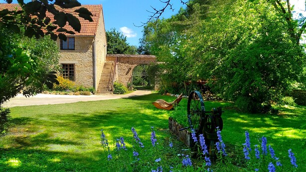 A stone building with a red-tiled roof, a stone staircase, and a garden with blue flowers and a windmill. - Jardin des Sens (Éguilly)