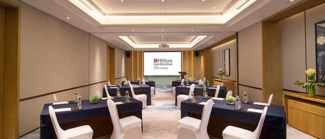 A conference room with a large screen displaying 'Hilton Garden Inn' branding, multiple tables with white chairs, and a carpeted floor.