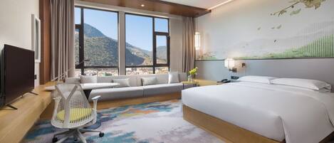 A hotel room with a large bed, a sofa, a desk with a chair, a TV, and a view of mountains.