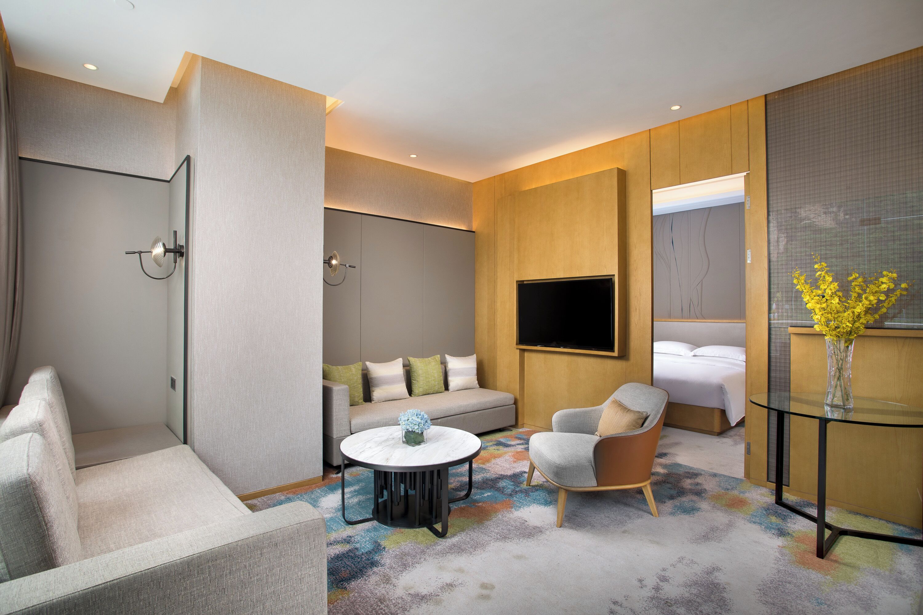 Una habitación de hotel moderna con sofá, sillón, una mesa redonda y televisor montado en la pared.