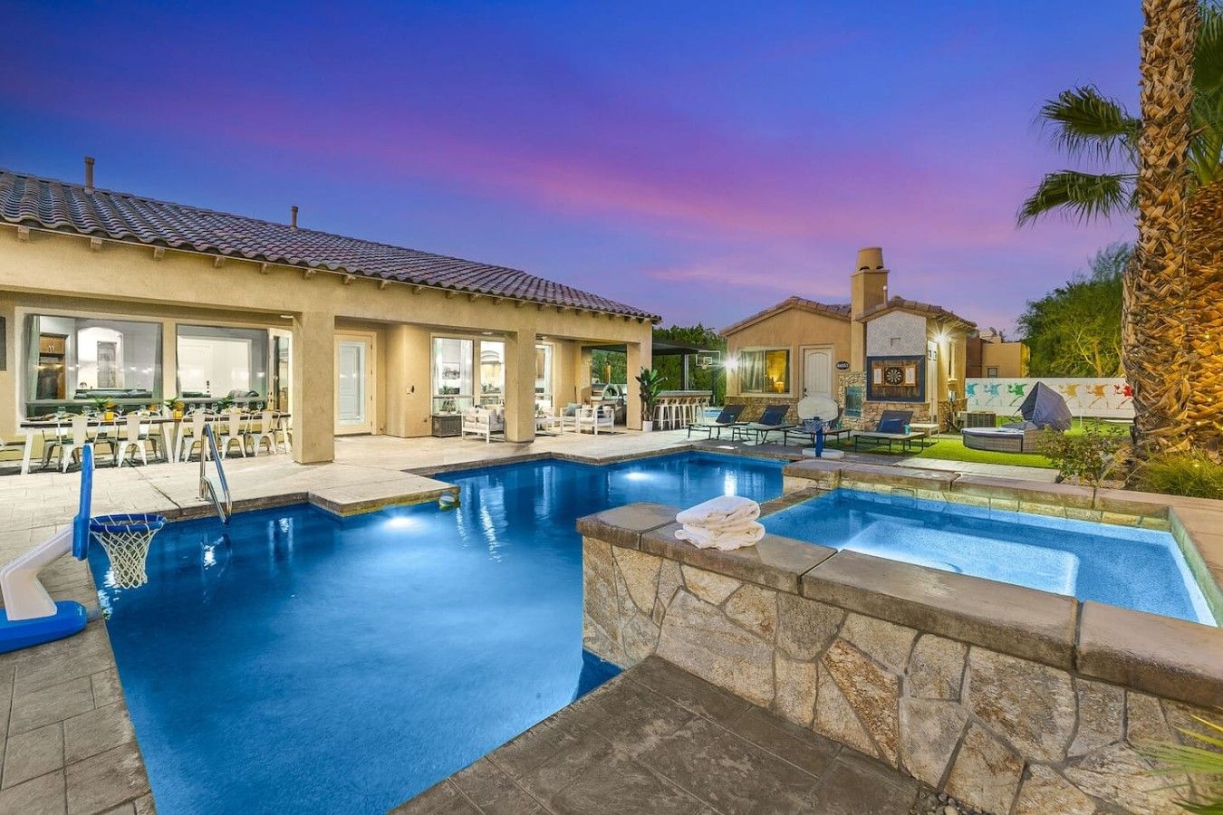 A swimming pool with a basketball hoop, lounge chairs, and a covered patio area.