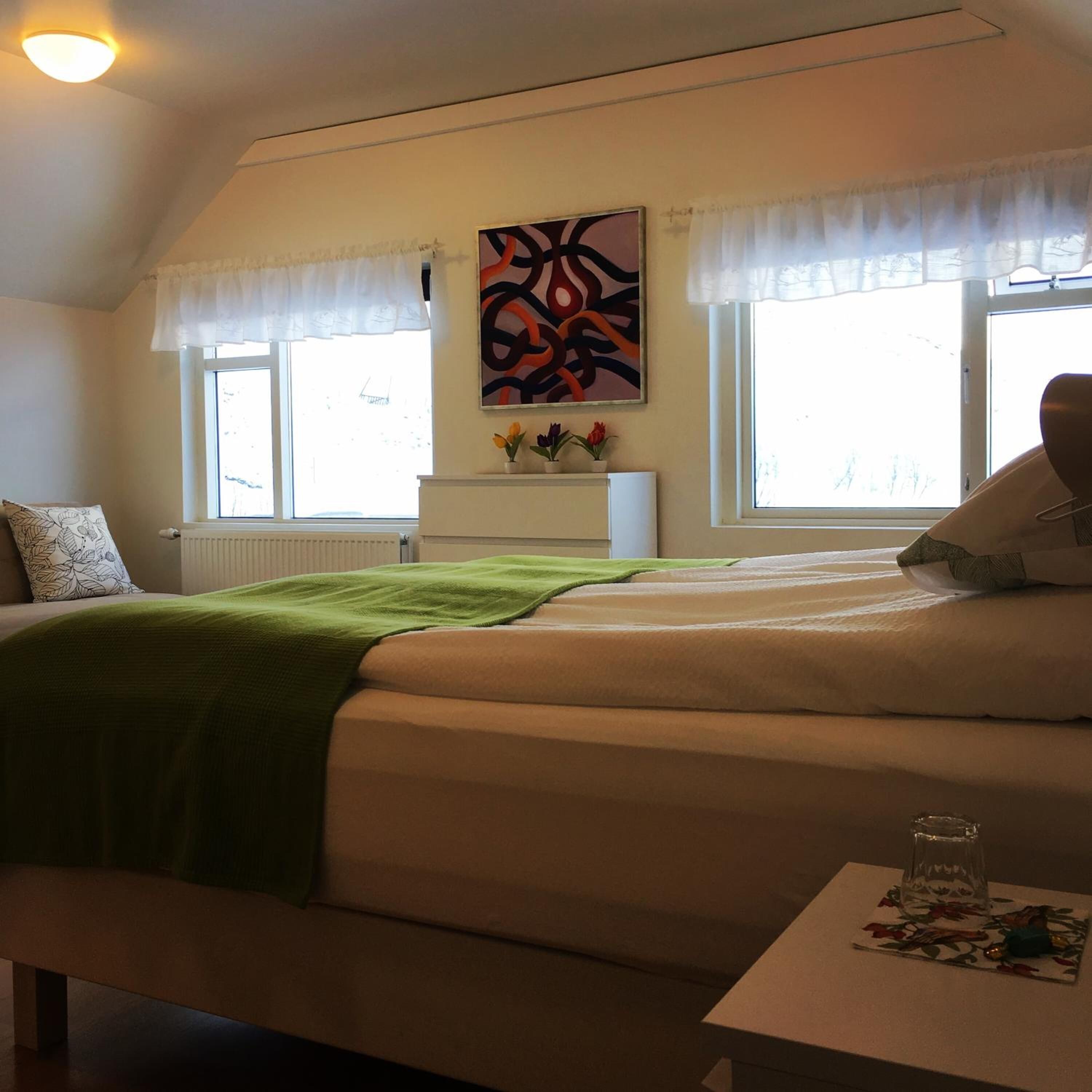 A bedroom with a bed, a bedside table, a radiator, a window with curtains, a wall-mounted painting, and a view of the outside.