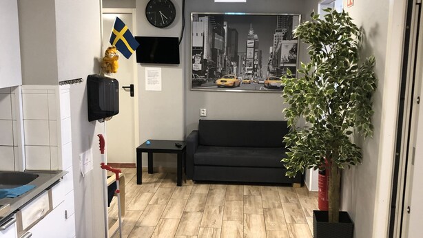 A reception area with a black sofa, a clock, a framed cityscape picture, a Swedish flag, a plant, and a trash bin. - Golden Hostel (Jarfalla)