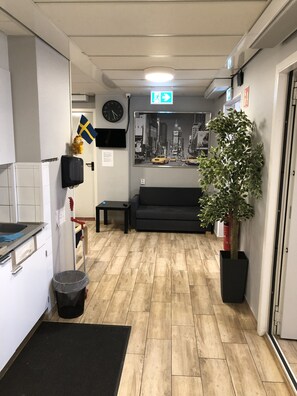 A reception area with a black sofa, a clock, a framed cityscape picture, a Swedish flag, a plant, and a trash bin. - Golden Hostel (Jarfalla)
