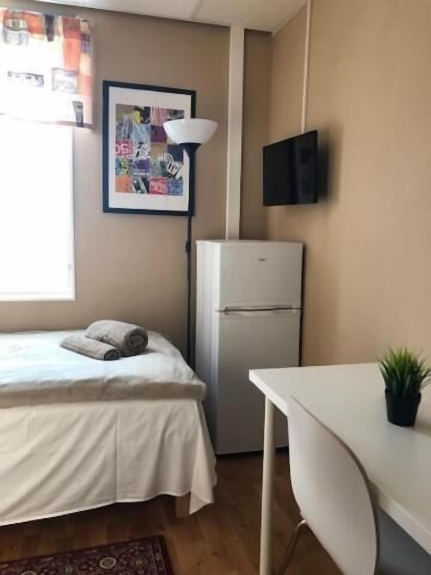 A bedroom with a bed, a desk, a refrigerator, a wall-mounted TV, and a window with curtains.
