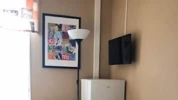 A bedroom with a bed, a desk, a refrigerator, a wall-mounted TV, and a window with curtains.