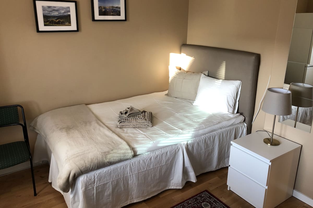 A neatly made bed with a white comforter and pillows, a small bedside table with a lamp, and two framed pictures on the wall.