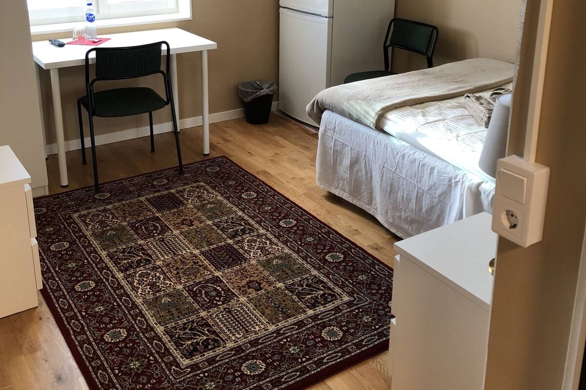 A small, well-lit room with a desk, chair, bed, and a refrigerator.