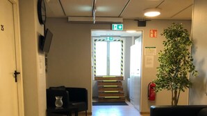 A hallway with a wooden floor, a black sofa, a small table with a vase, a plant, and a staircase leading to an exit. - Golden Hostel (Jarfalla)