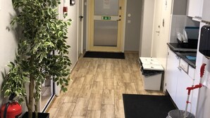 A hallway with a wooden floor, a potted plant, a fire extinguisher, a trash bin, and a kitchen area with cabinets and a sink. - Golden Hostel (Jarfalla)