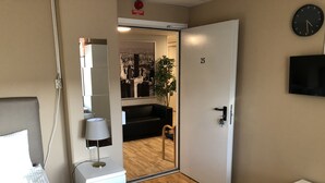 A modern bedroom with a bed, bedside table, lamp, and a small white dresser. A mirror is mounted on the wall, and a clock is visible on the wall. - Golden Hostel (Jarfalla)