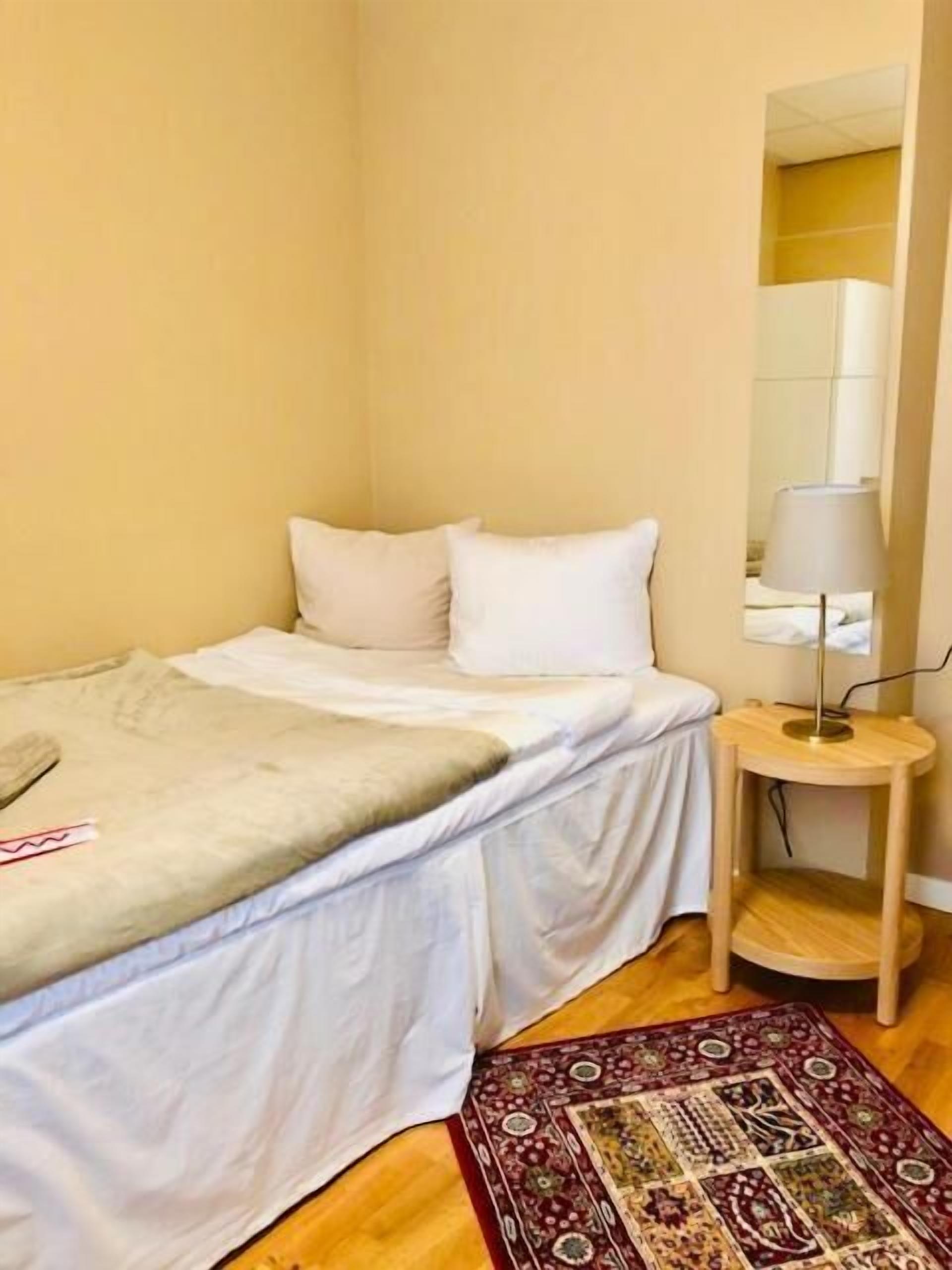 A small, neatly made bed with white bedding and a beige blanket, a small wooden side table with a lamp, and a patterned rug on a wooden floor.