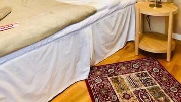 Una cama pequeña y bien arreglada con ropa blanca y una manta color beige, una mesita de madera con una lámpara, y una alfombra estampada sobre un piso de madera.