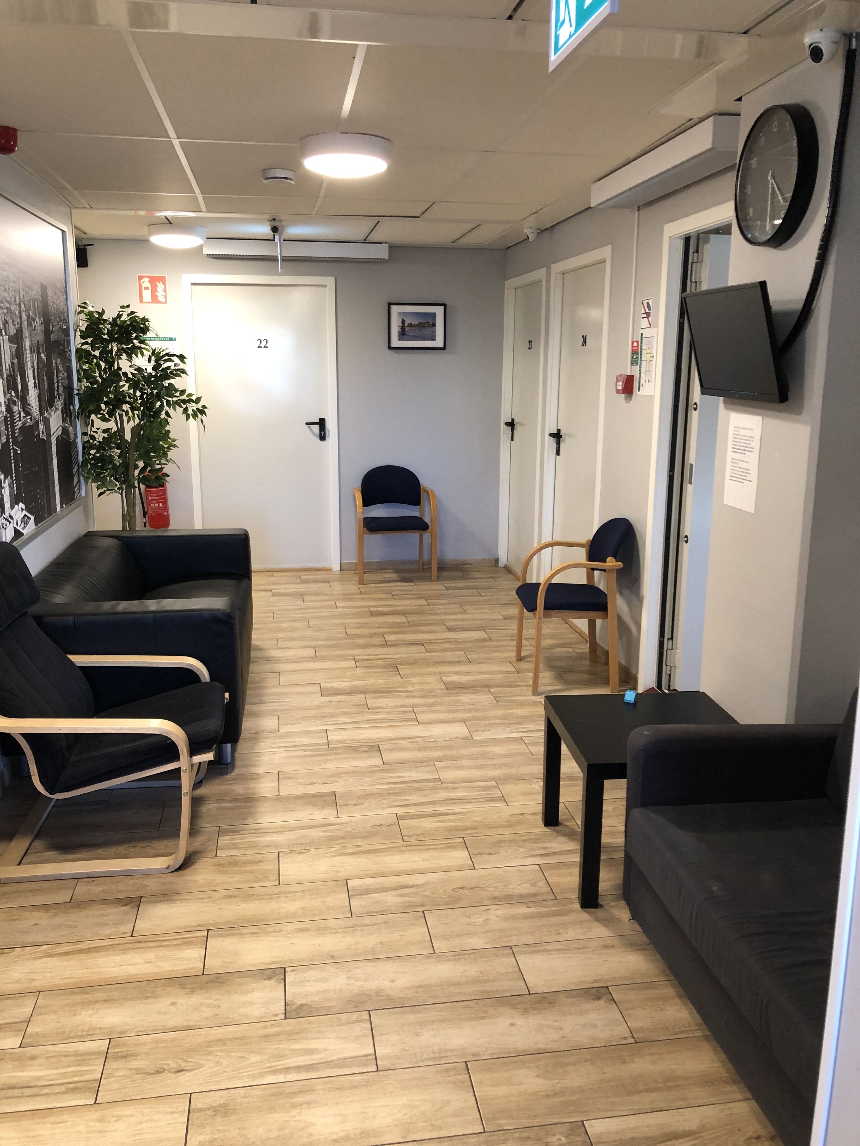 A reception area with a wooden herringbone floor, black leather sofas, a wooden chair, a small table, and a wall-mounted clock.