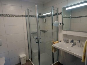 Un baño moderno con ducha envuelta en vidrio, un lavamanos blanco con un espejo arriba y suelo de baldosas.