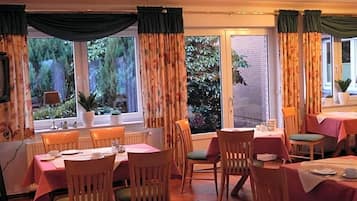 A dining area with tables and chairs, large windows with curtains, and a view of greenery outside.