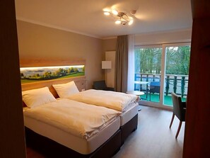 Habitación de hotel con cama, escritorio, silla, ventana con vistas a vegetación y balcón con mobiliario exterior.