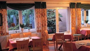 A dining area with tables and chairs, large windows with curtains, and a view of greenery outside.