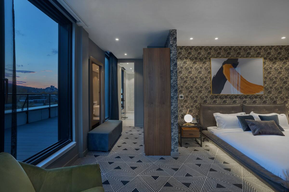 A modern hotel room with a large window, a bed with multiple pillows, a nightstand with a lamp, and a geometric patterned floor.