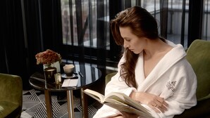 Eine Frau sitzt auf einem grünen Sofa und liest ein Buch in einem modernen Raum mit einem gemusterten Boden, einem kleinen Tisch mit Blumenvase und Blick auf die Stadt durch große Fenster.