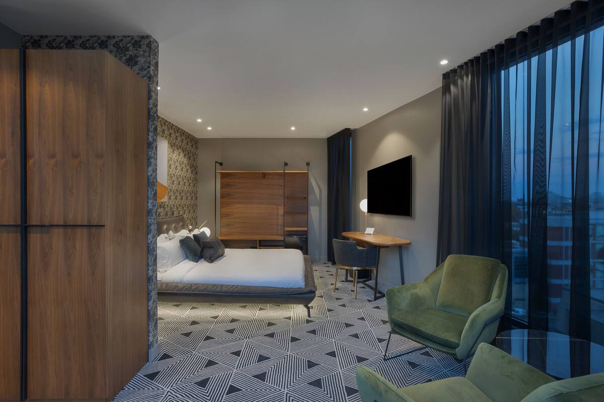 A modern hotel room with a bed, a desk, two armchairs, and a large window with curtains.