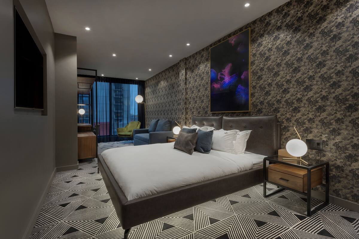 A modern bedroom with a large bed, a nightstand with a lamp, a wall-mounted TV, and a window with curtains.