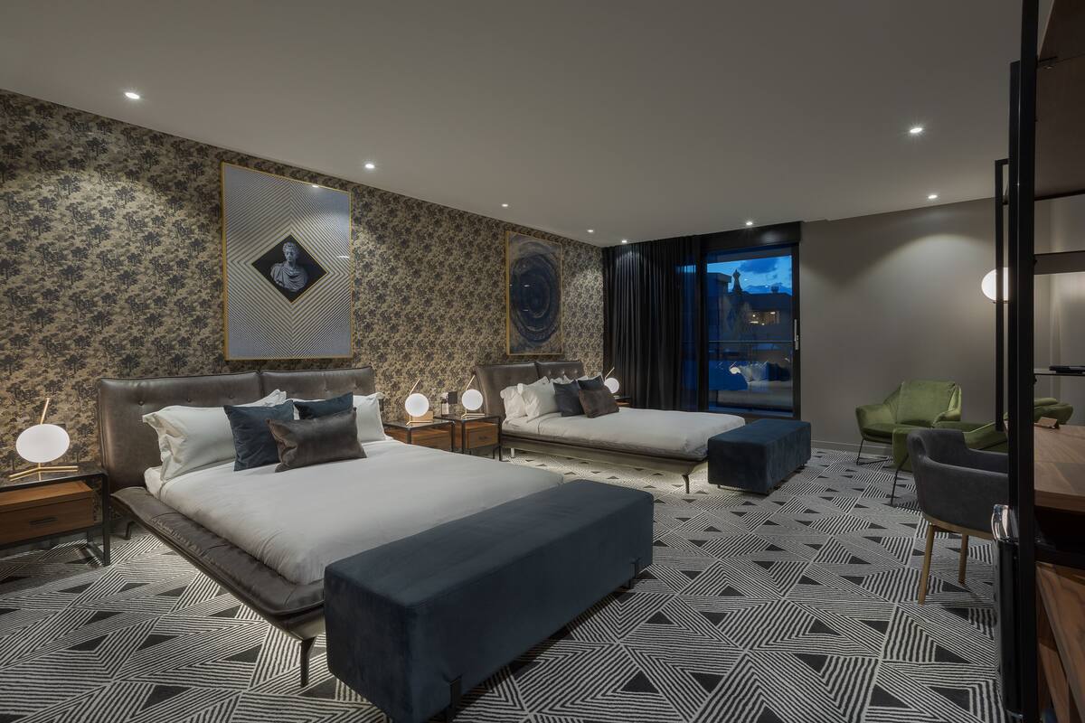 A modern hotel room with two beds, a large window, and a view of the cityscape.
