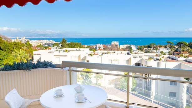 A balcony with a white table and chairs, overlooking a coastal cityscape with buildings and the sea.
