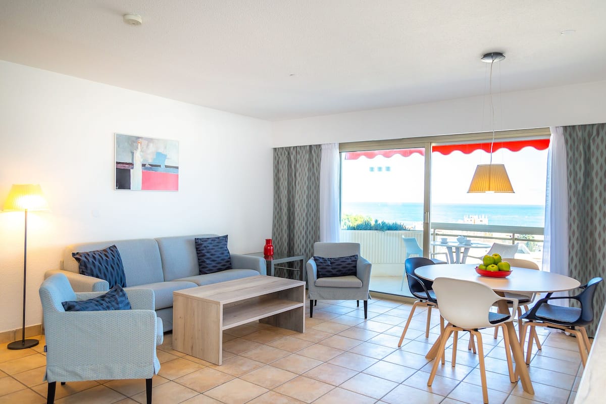A modern living room with a sofa, coffee table, and dining area. There is a painting on the wall, a lamp, and a view of the sea through large windows.