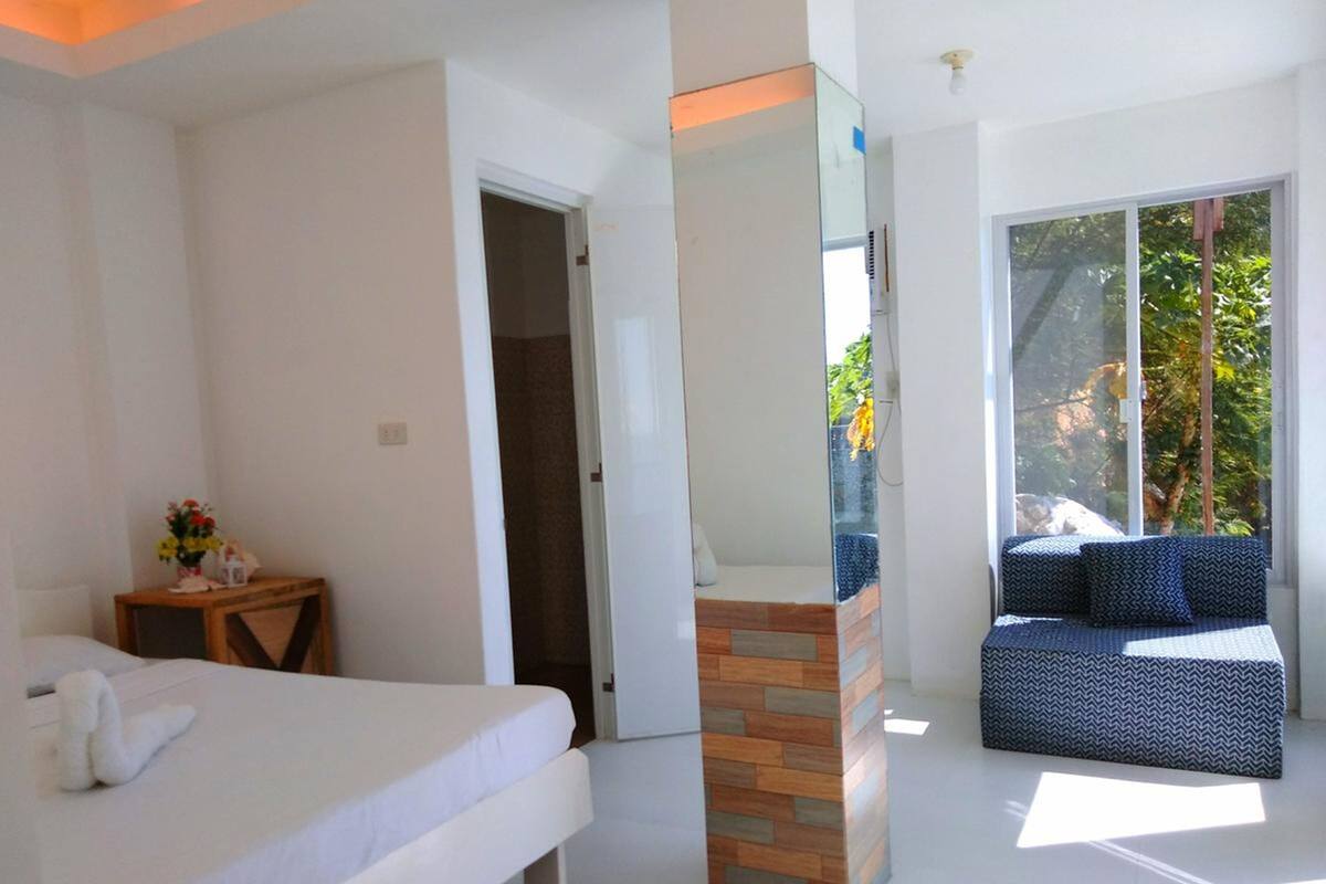 Ein modernes Schlafzimmer mit Bett, einem kleinen Tisch mit Blumenvase, einer Sitzecke mit gemusterten Kissen und einem großen Fenster mit Blick ins Grüne.